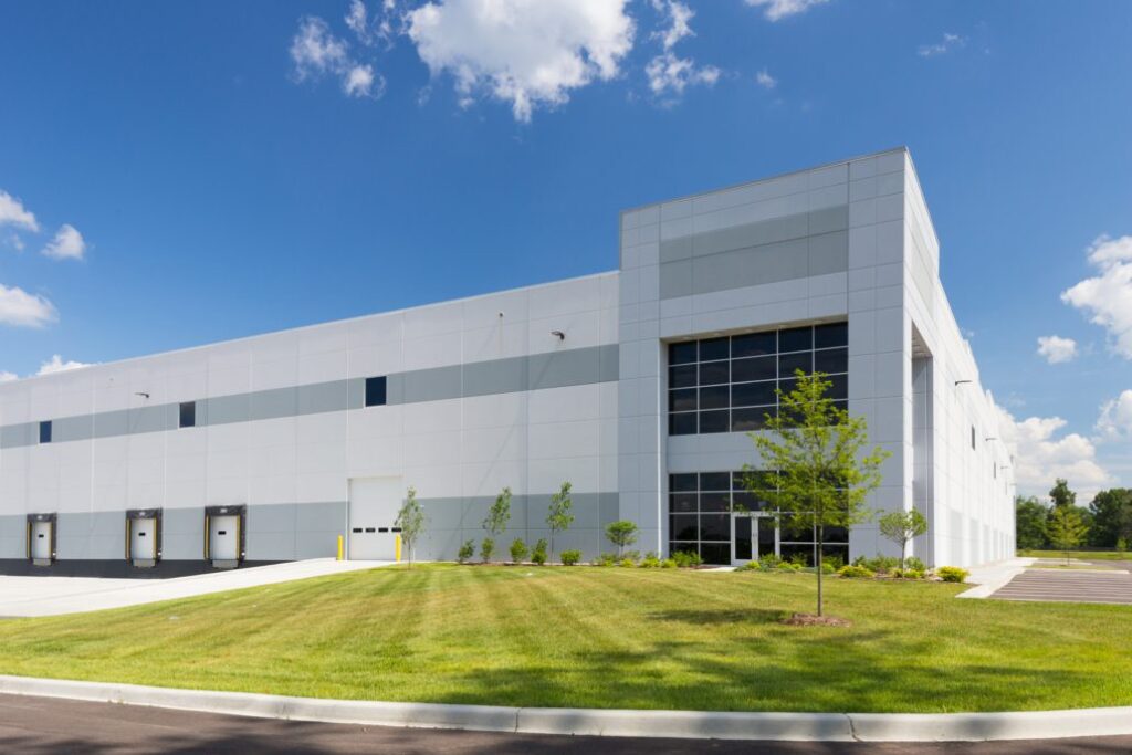Large white industrial warehouse with multiple loading dock doors, glass entrance section, landscaped lawn, and a clear blue sky above.