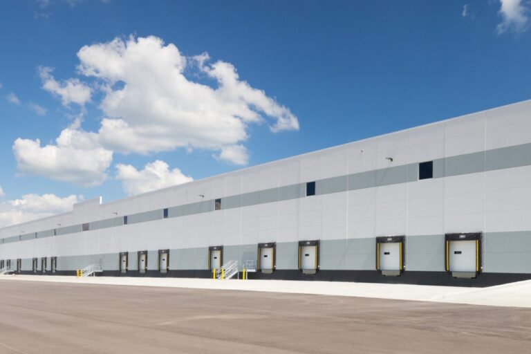 Long industrial warehouse with rows of loading dock doors, light gray paneling, and a wide paved lot under a bright blue sky with clouds.