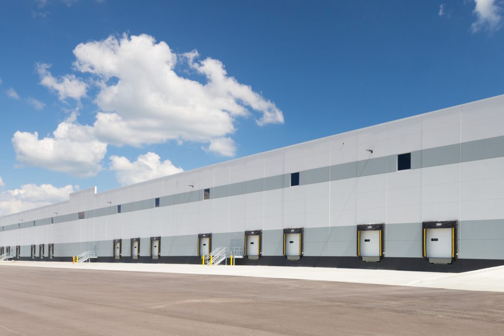 Long industrial warehouse with rows of loading dock doors, light gray paneling, and a wide paved lot under a bright blue sky with clouds.