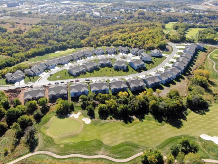 Curved rows of houses with gray roofs border a golf course with sand traps and green fairways, surrounded by trees and open landscape.