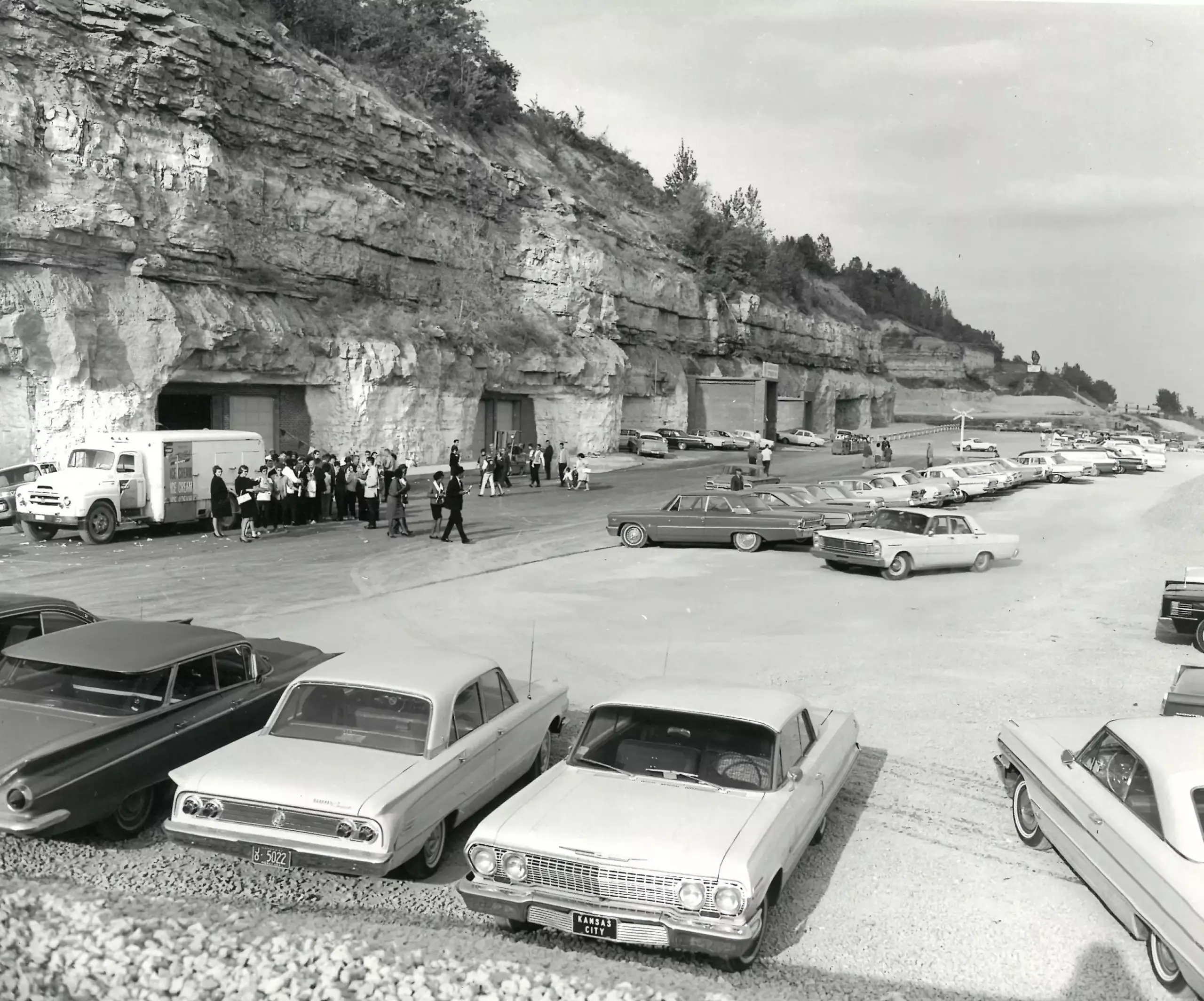 Black-and-white photo of 1960s cars parked near cave entrances in a rock cliff, with a crowd gathered by a truck and building doors.