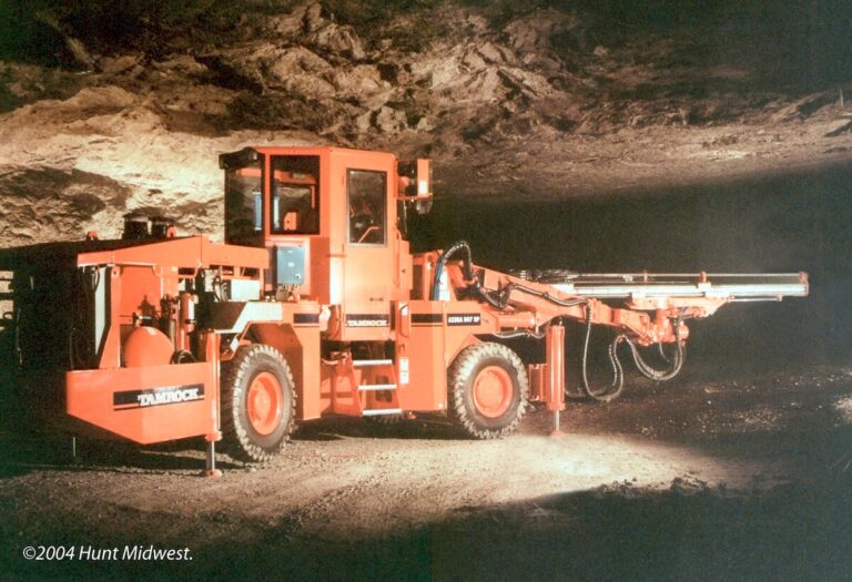 Bright orange Tamrock drilling machine with extendable arm parked in an underground limestone mine, illuminated against the rock walls and ceiling.