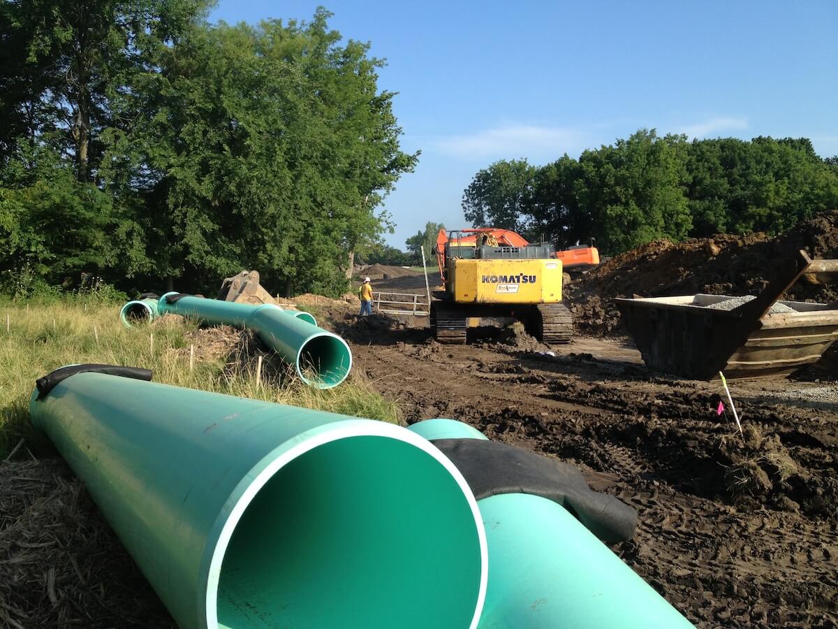 Large green utility pipes lie in the grass beside a muddy worksite where a yellow Komatsu excavator is operating. A worker in safety gear stands near the equipment, with piles of dirt and gravel bins nearby, surrounded by trees under a clear sky.