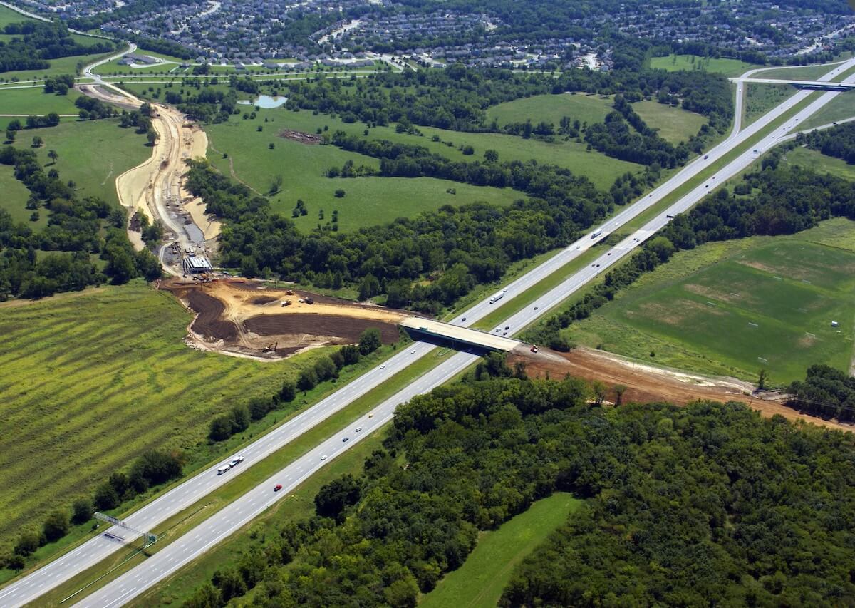 Wide aerial shot showing an active construction site near a bridge overpass, with cleared land, heavy equipment, and adjacent highways surrounded by green fields and residential areas.