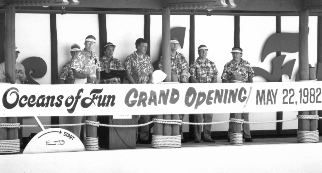 Black-and-white photo of the May 22, 1982 Oceans of Fun water park grand opening. A group of men in Hawaiian shirts and visors stand behind a banner announcing the event.