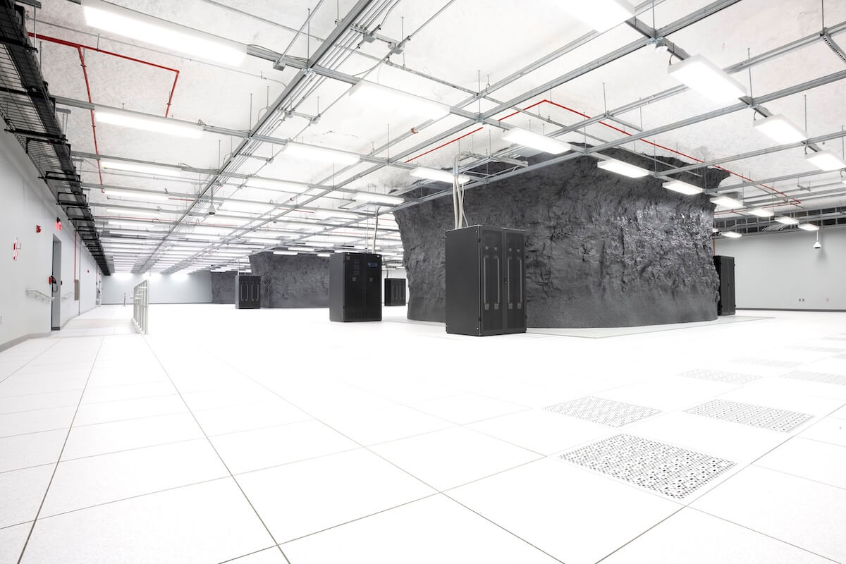 A bright, spacious data center with white tile flooring and overhead cable trays features black server cabinets placed around large exposed rock columns, blending natural stone with modern infrastructure.