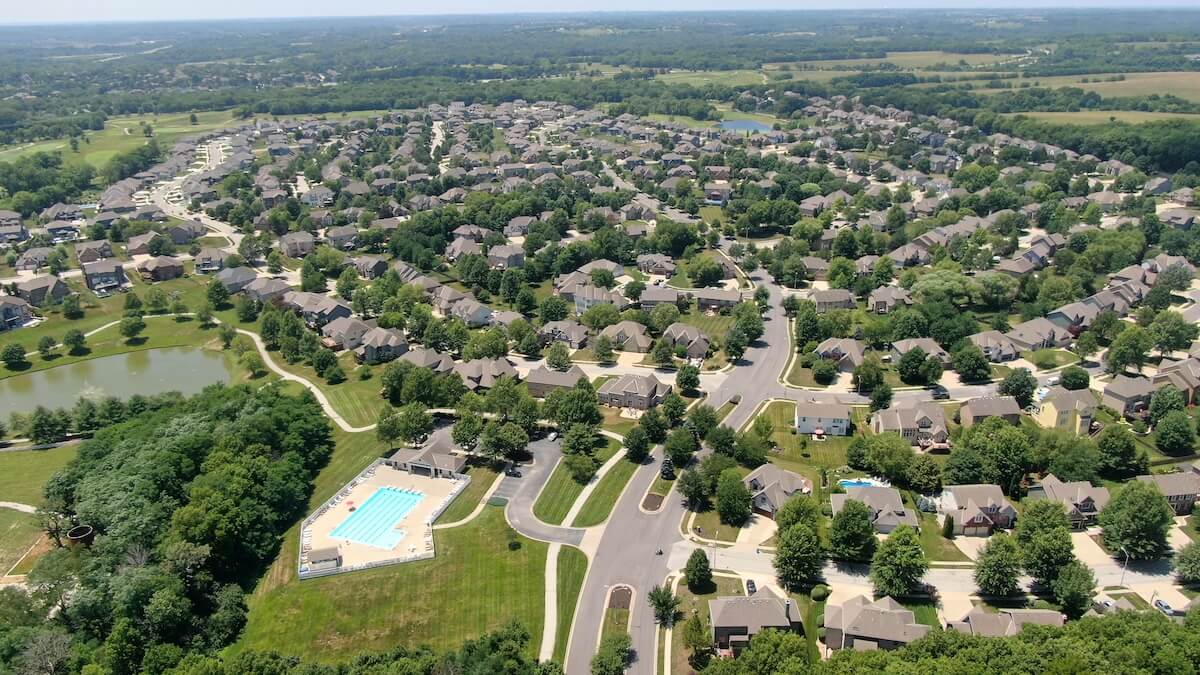 Expansive housing development with winding streets, numerous homes, a community swimming pool, and small lakes surrounded by greenery.