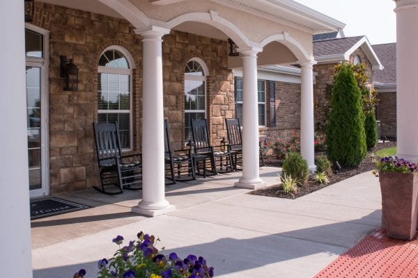 Stone exterior building with white columns, lined rocking chairs, potted flowers, and landscaped walkway creating a welcoming outdoor seating area.
