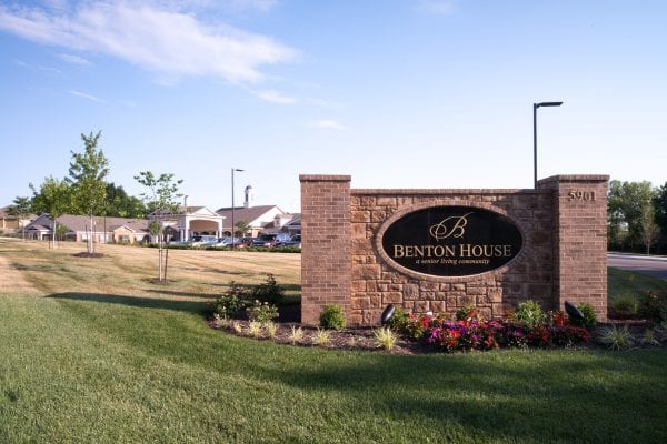 Brick monument sign with landscaping and flowers marking the entrance to Benton House, a senior living community.