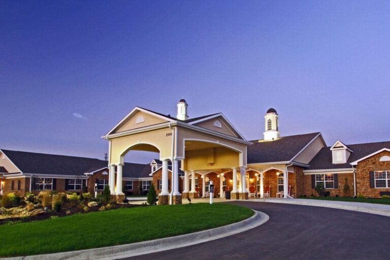 Evening view of senior living facility featuring a large covered entry with columns, warm lighting, brick exterior, and landscaped driveway.