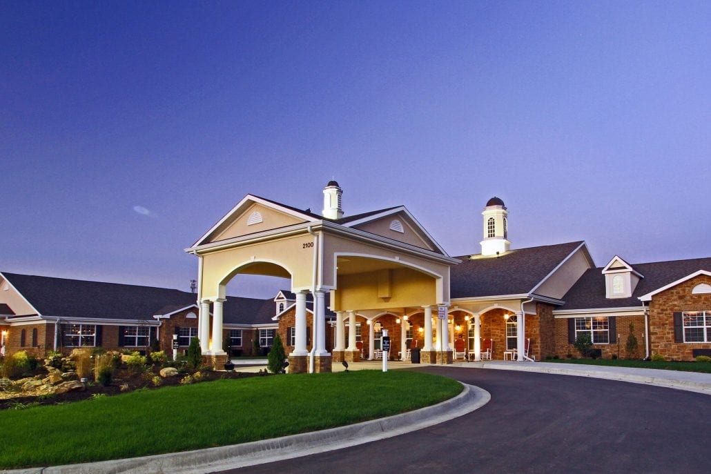 Evening view of senior living facility featuring a large covered entry with columns, warm lighting, brick exterior, and landscaped driveway.