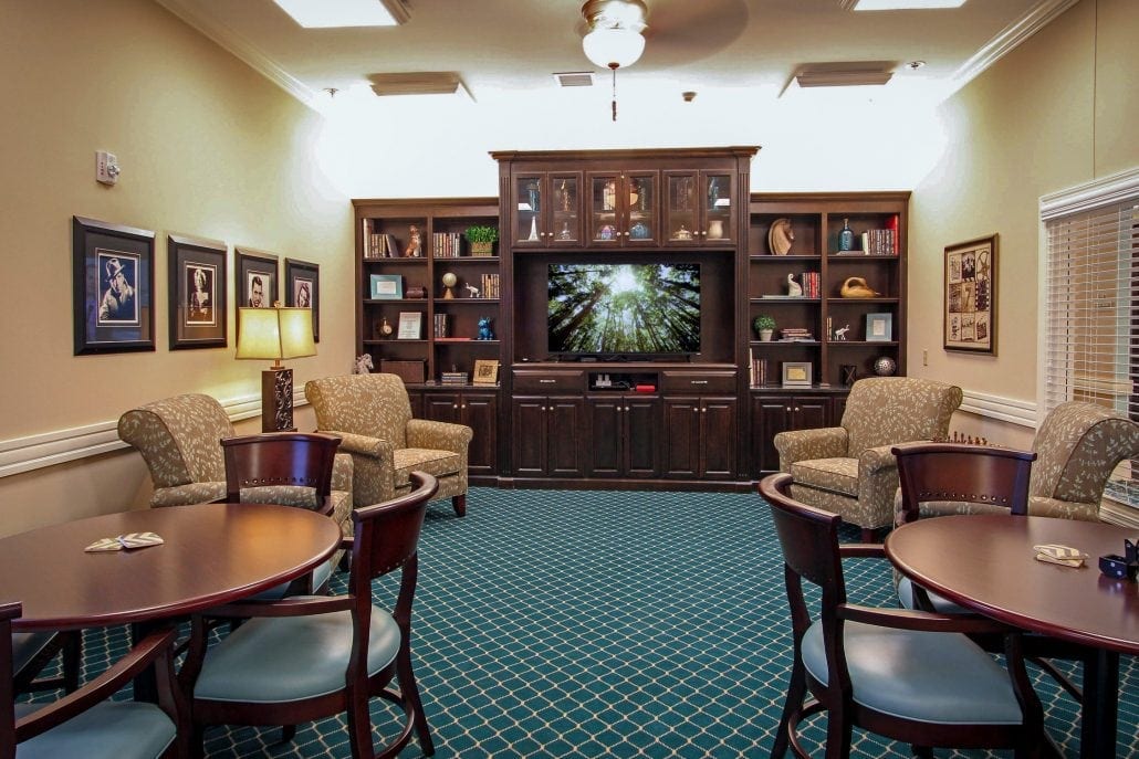 Cozy library and media lounge with dark wood built-in shelving, a flat-screen TV, patterned teal carpet, upholstered armchairs, and round wooden tables with chairs for reading, games, or small gatherings.