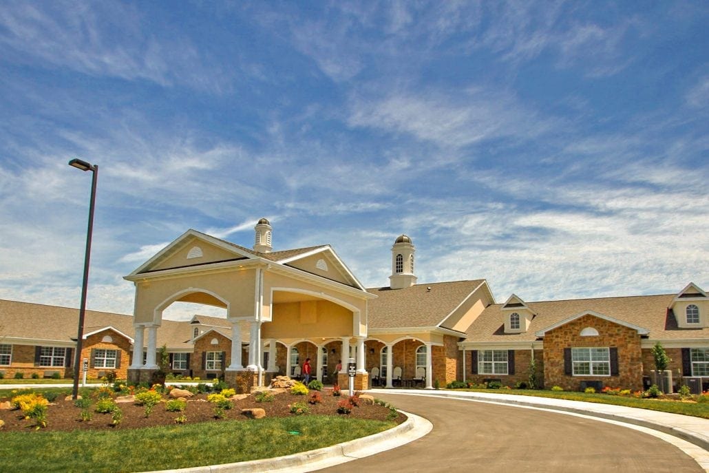 Front view of senior living facility with covered entryway, brick and siding exterior, landscaped garden, and circular driveway.