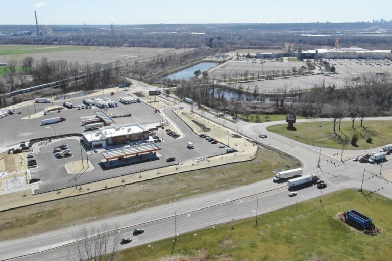 Large fueling station with multiple semi-trucks parked, surrounded by roads, parking lots, and nearby industrial and retail areas.