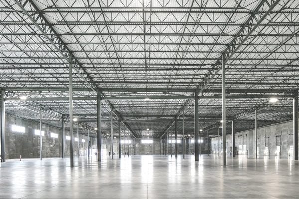 Vast empty warehouse interior with polished concrete floors, high ceilings, and rows of steel support beams. The ceiling is constructed with a metal truss system and skylights allowing natural light to filter in. The space appears industrial, open, and ready for storage or manufacturing use.