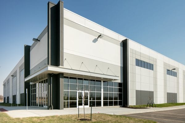 Modern industrial warehouse exterior with a long white facade accented by dark vertical columns. The building features large glass windows and double entry doors with metal awnings above. A clean asphalt parking lot with marked spaces is visible in the foreground under a clear blue sky.