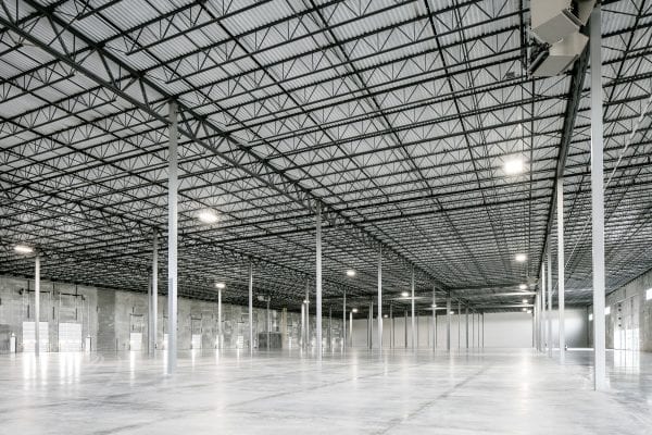 Spacious empty warehouse interior with smooth concrete flooring, tall ceilings, and rows of steel support columns. The ceiling features a grid of exposed metal trusses with industrial lighting fixtures. Large roll-up doors and windows line the concrete walls, allowing natural and artificial light to illuminate the vast open space.