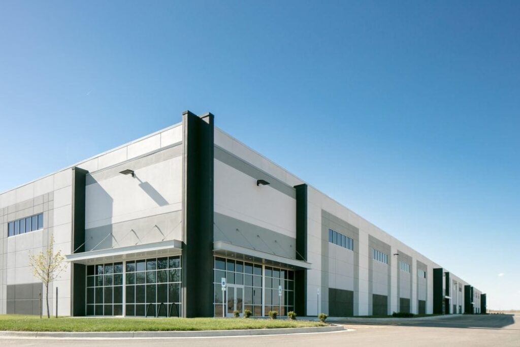 Large white and gray warehouse with tall black columns, glass entrance, and rows of windows under a clear blue sky.