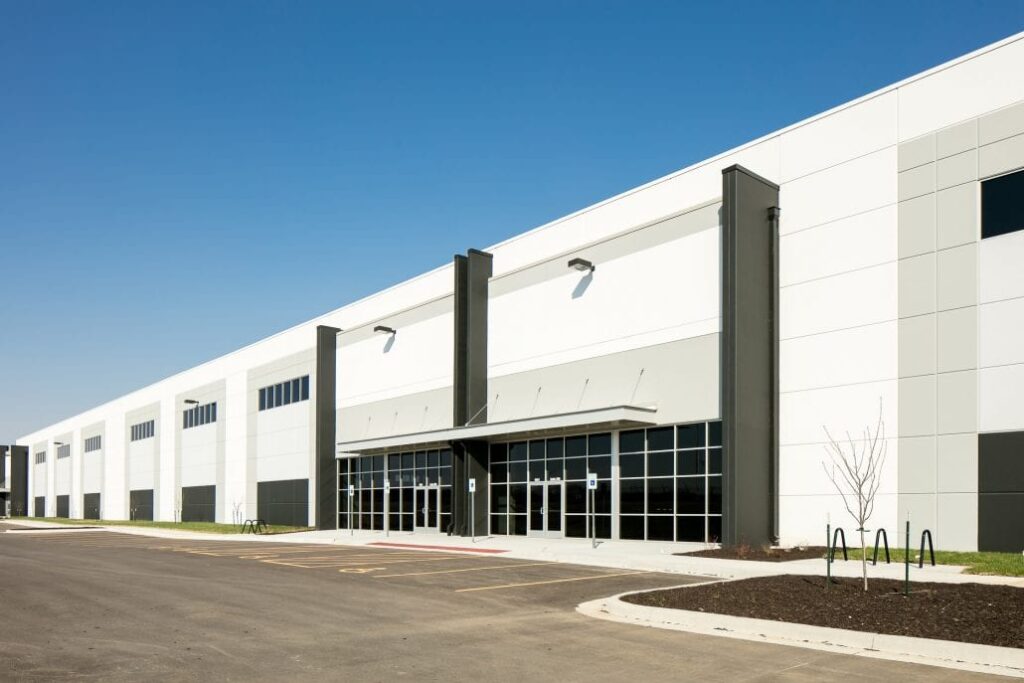 Long white warehouse building with black columns, glass-fronted entrance, and parking area under a clear blue sky.