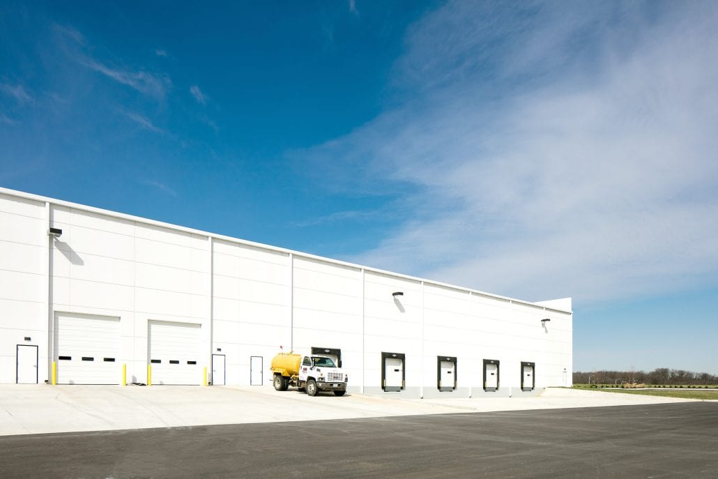 Wide view of a modern white warehouse exterior with multiple loading dock doors and a yellow water truck parked in front.