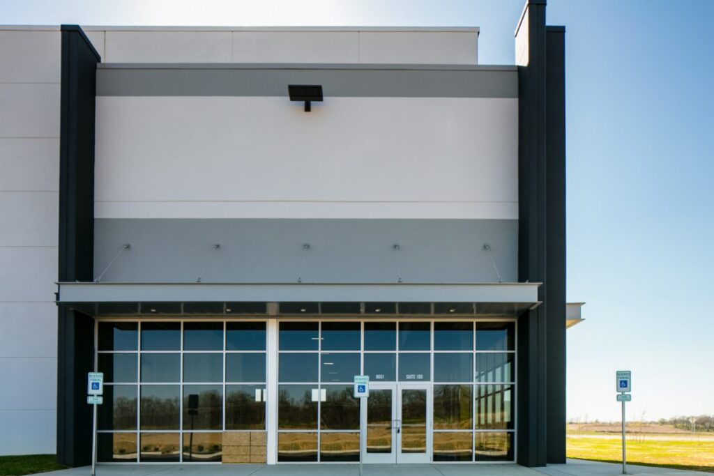 Modern warehouse entrance with tall glass windows, double doors, and handicap parking signs under a flat canopy.