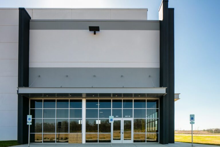 Modern warehouse entrance with tall glass windows, double doors, and handicap parking signs under a flat canopy.