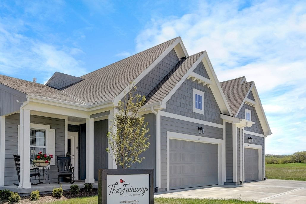 Gray single-story home with pitched roofs, white trim, front porch seating, and a garage, with a sign reading 'The Fairways.'