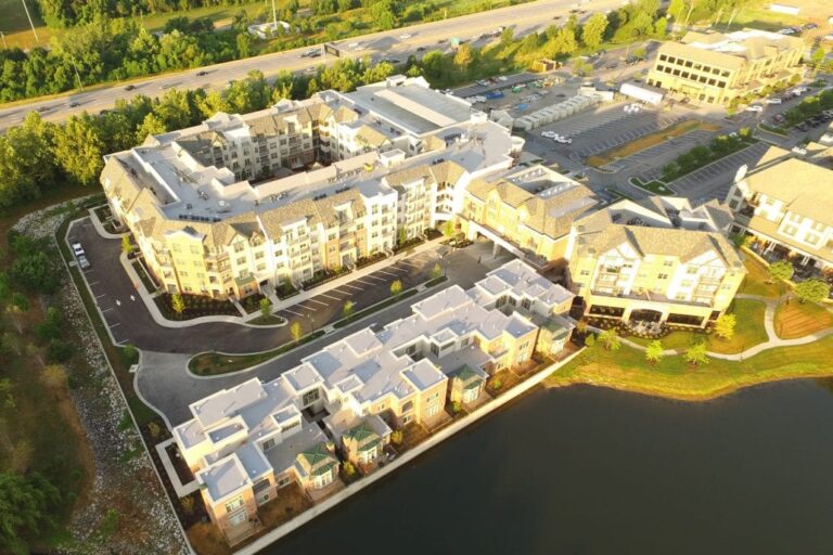 Drone shot of a large multi-story apartment community with varied rooflines, parking areas, and landscaped paths along a waterfront.