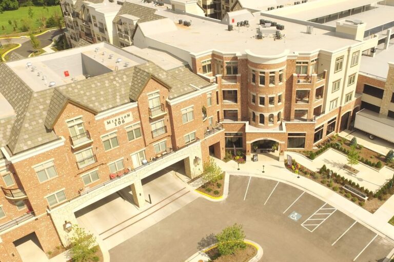 Brick and stone multi-story apartment complex with balconies, arched entryway, landscaped courtyard, and adjacent parking area.