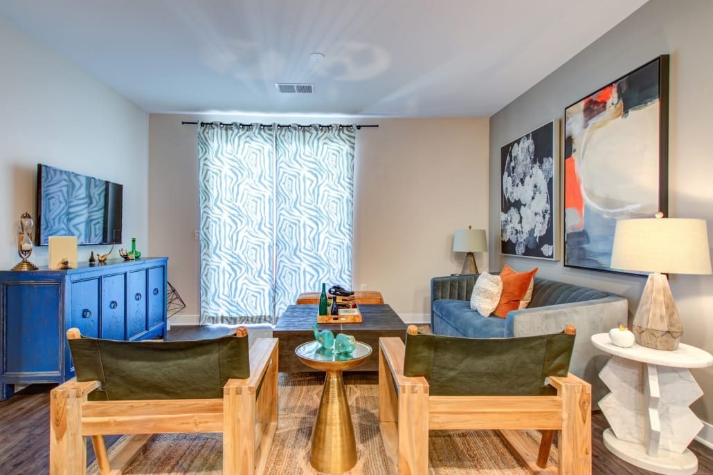 Bright living room featuring a blue sofa, wooden chairs, patterned curtains, abstract wall art, and a flat-screen TV on a blue console.