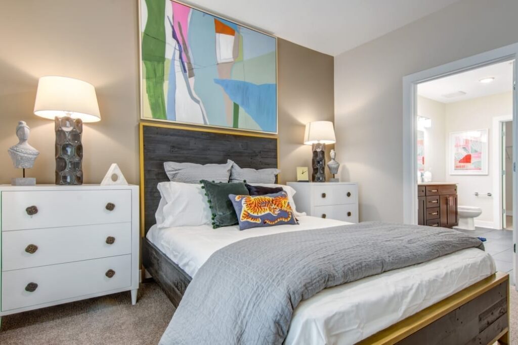 Cozy bedroom with a wood-frame bed, gray bedding, colorful pillows, abstract artwork above the headboard, and an adjoining bathroom.