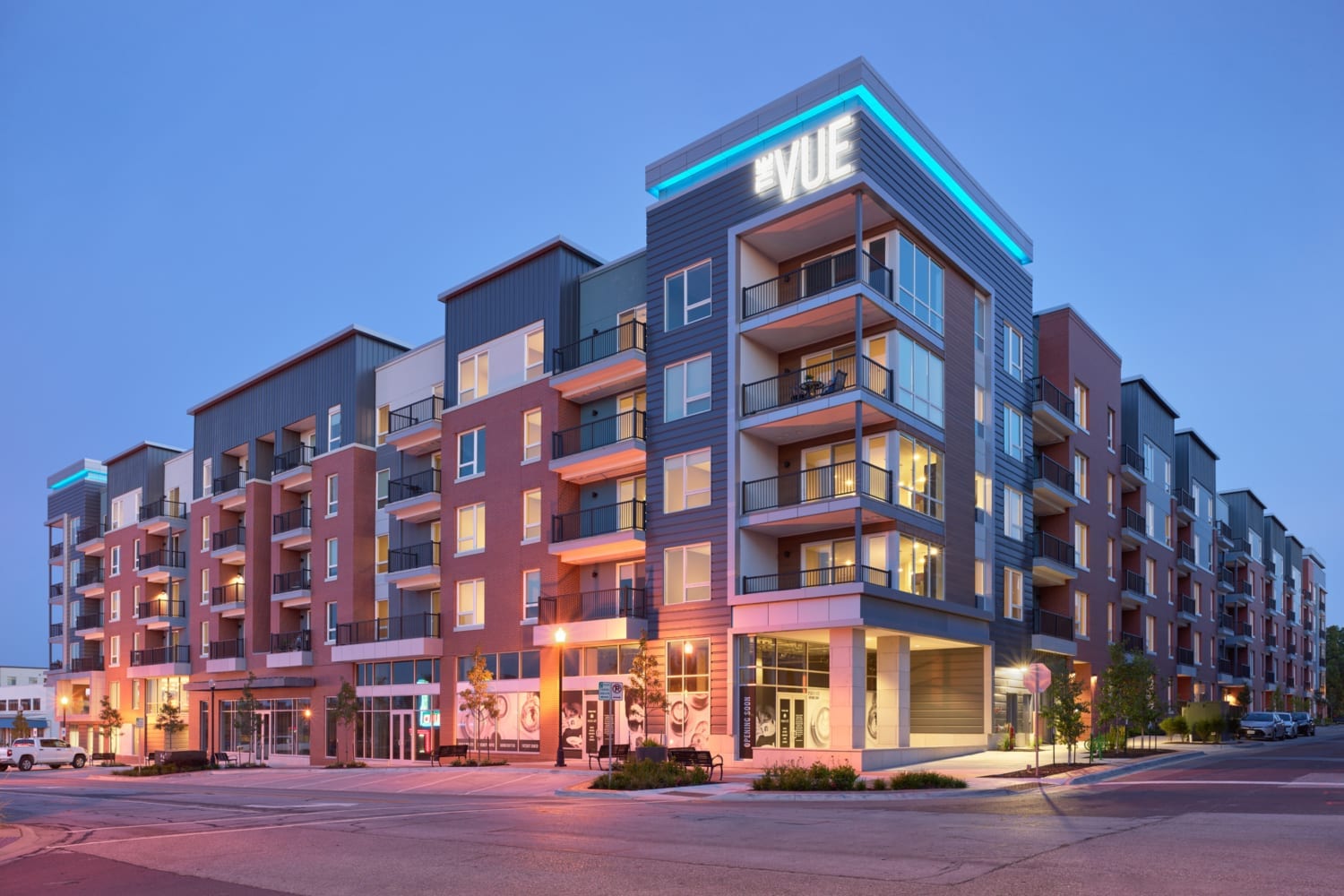 Street view of The Vue apartments, a multi-story mixed-use building with balconies, large windows, and illuminated signage at twilight.