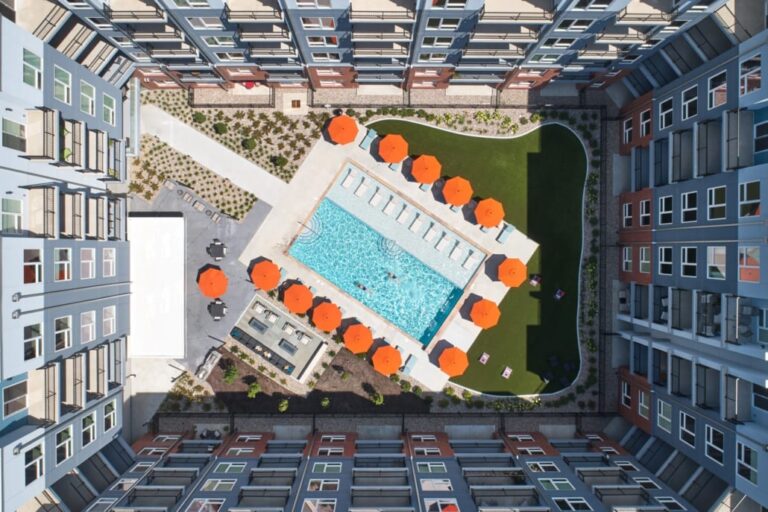 Rectangular swimming pool surrounded by lounge chairs and bright orange umbrellas, framed by multi-story apartment buildings.