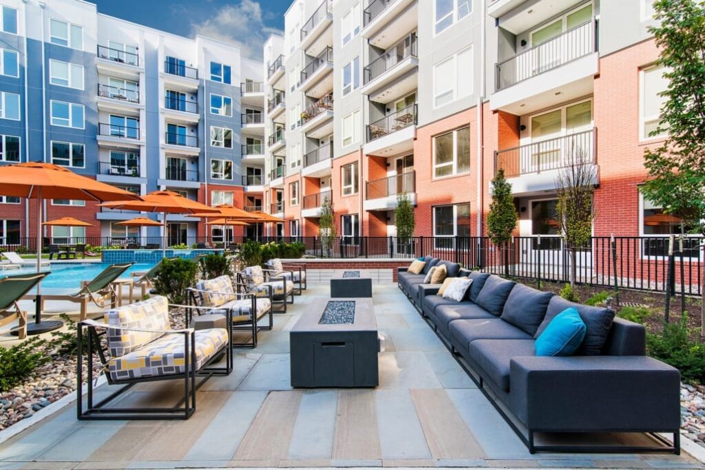 Courtyard with cushioned chairs, long sofas, fire pits, orange umbrellas, and poolside loungers surrounded by modern apartments.