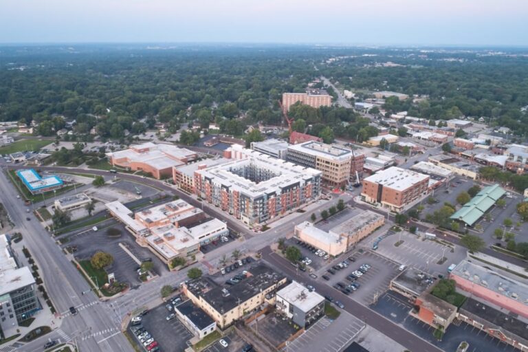 Drone view showing The Vue apartment complex with central courtyard pool, surrounded by shops, streets, and nearby residential areas.