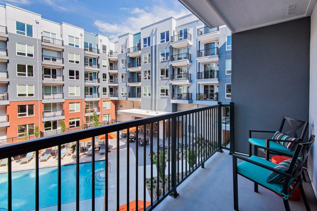 Private balcony with chairs and cushions overlooking a courtyard pool, lounge areas, and multi-story apartment building.
