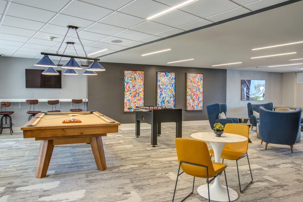 Modern game room featuring a pool table, foosball table, colorful wall art, TV, and seating with yellow and blue chairs.