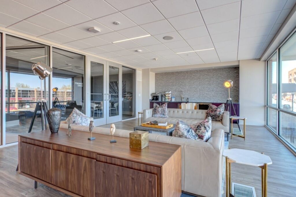 Bright lounge with cream sofas, patterned pillows, wood cabinet, decorative sculptures, and a coffee bar near large windows.