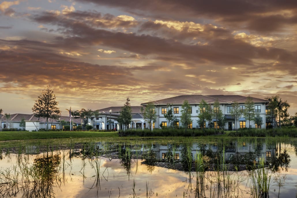 Scenic view of a senior living community building at sunset, with warm lights glowing from the windows. The two-story structure has a red-tiled roof and is surrounded by trees and greenery. In the foreground, a calm pond reflects the building and the dramatic sky filled with golden clouds.