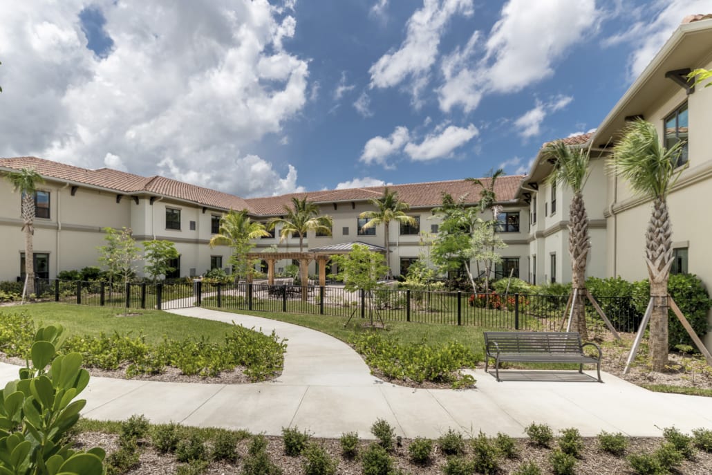 A landscaped courtyard surrounded by a two-story cream-colored building with a red-tiled roof. Palm trees, shrubs, and flowering plants line the area, along with a paved walkway curving toward a central pergola and outdoor seating space. A black metal fence encloses the patio, and a bench sits near the path in the foreground. The sky above is bright with large white clouds scattered across a vivid blue backdrop.