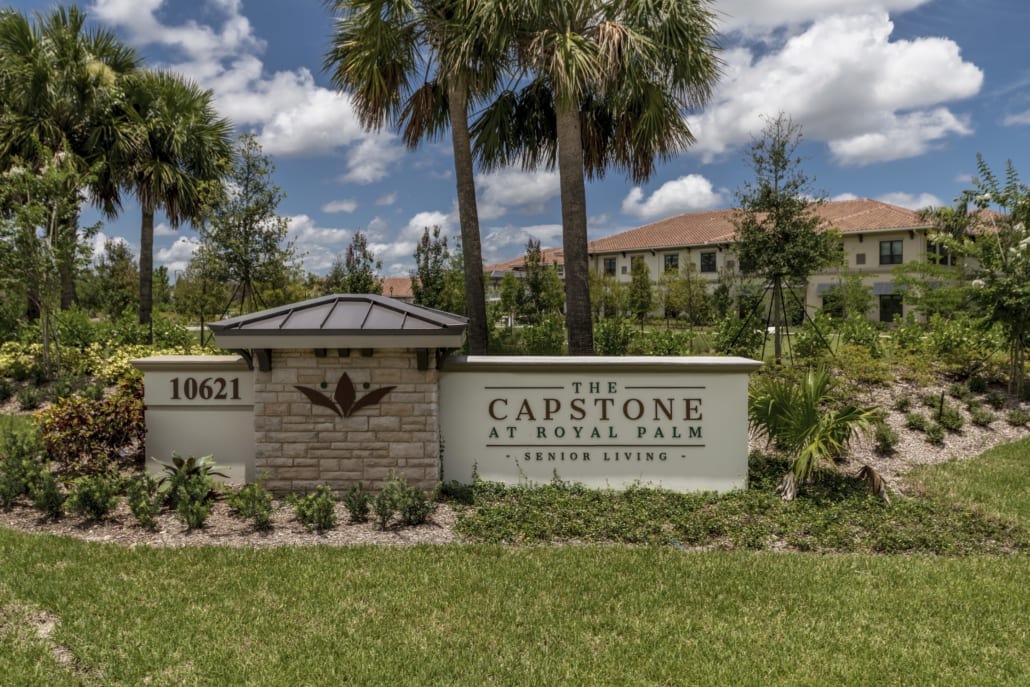Stone and stucco entrance sign for The Capstone at Royal Palm Senior Living with palm trees and landscaped greenery.