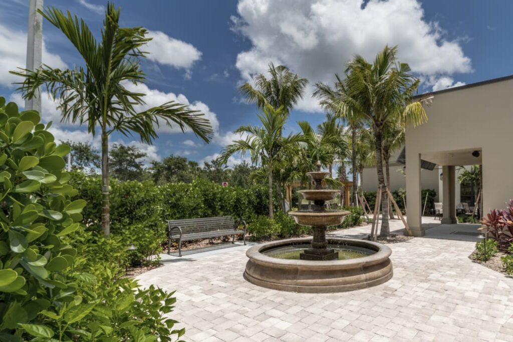 Stone courtyard featuring a tiered fountain, palm trees, greenery, and a metal bench under a sunny sky.