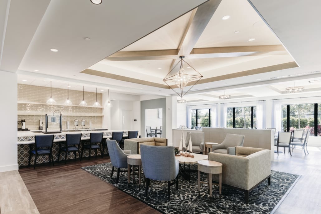 Bright lounge with modern chandeliers, cozy seating on a patterned rug, and a bar area with high-backed chairs and pendant lighting.