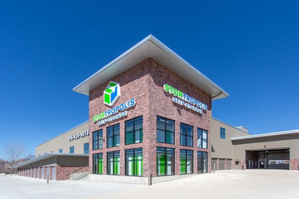 Modern brick and metal self-storage building with large windows, green doors, and 'Stortropolis Self-Storage' signage against a clear blue sky.