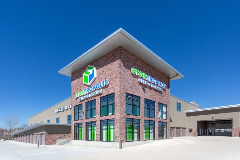 Modern brick and metal self-storage building with large windows, green doors, and 'Stortropolis Self-Storage' signage against a clear blue sky.