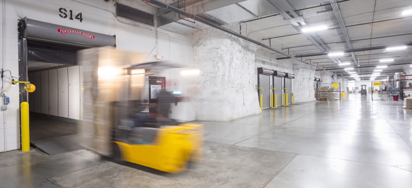 Underground warehouse with concrete floors, multiple loading dock doors, and a yellow forklift moving a pallet into a bay.