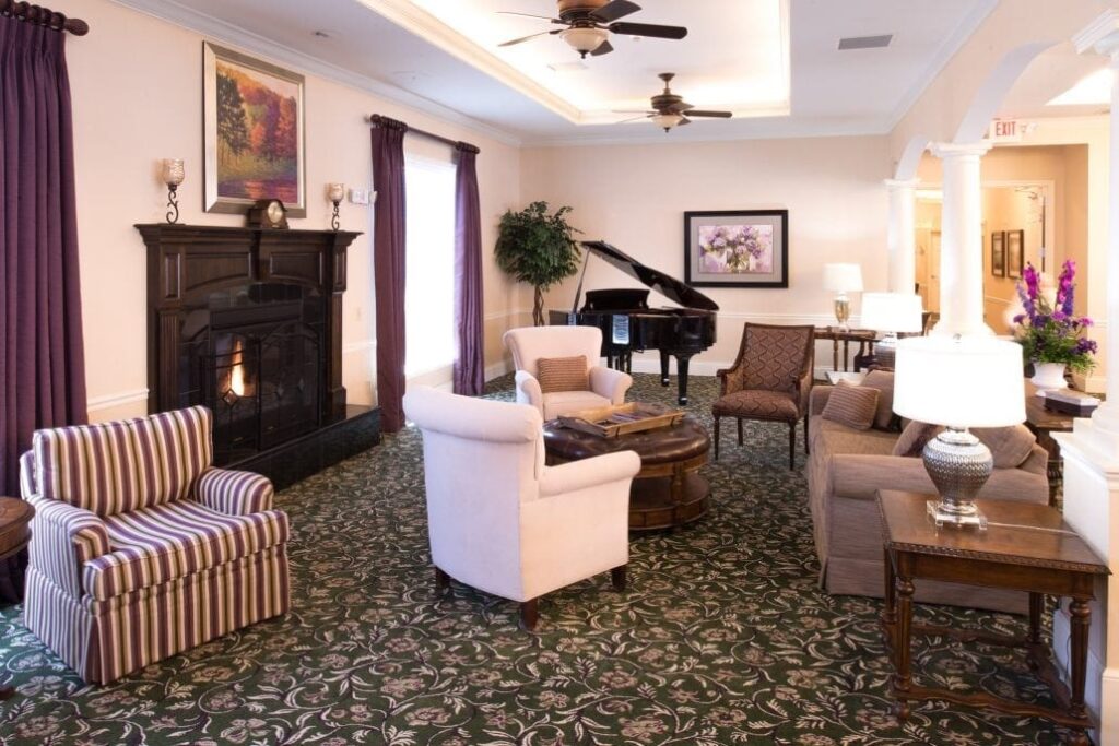 Elegant lounge featuring patterned carpet, upholstered chairs, sofas, a fireplace, grand piano, ceiling fans, and framed artwork.