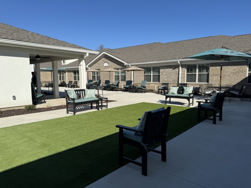 Open courtyard area with artificial grass, cushioned benches, lounge chairs, umbrellas, and brick building backdrop under a clear blue sky.