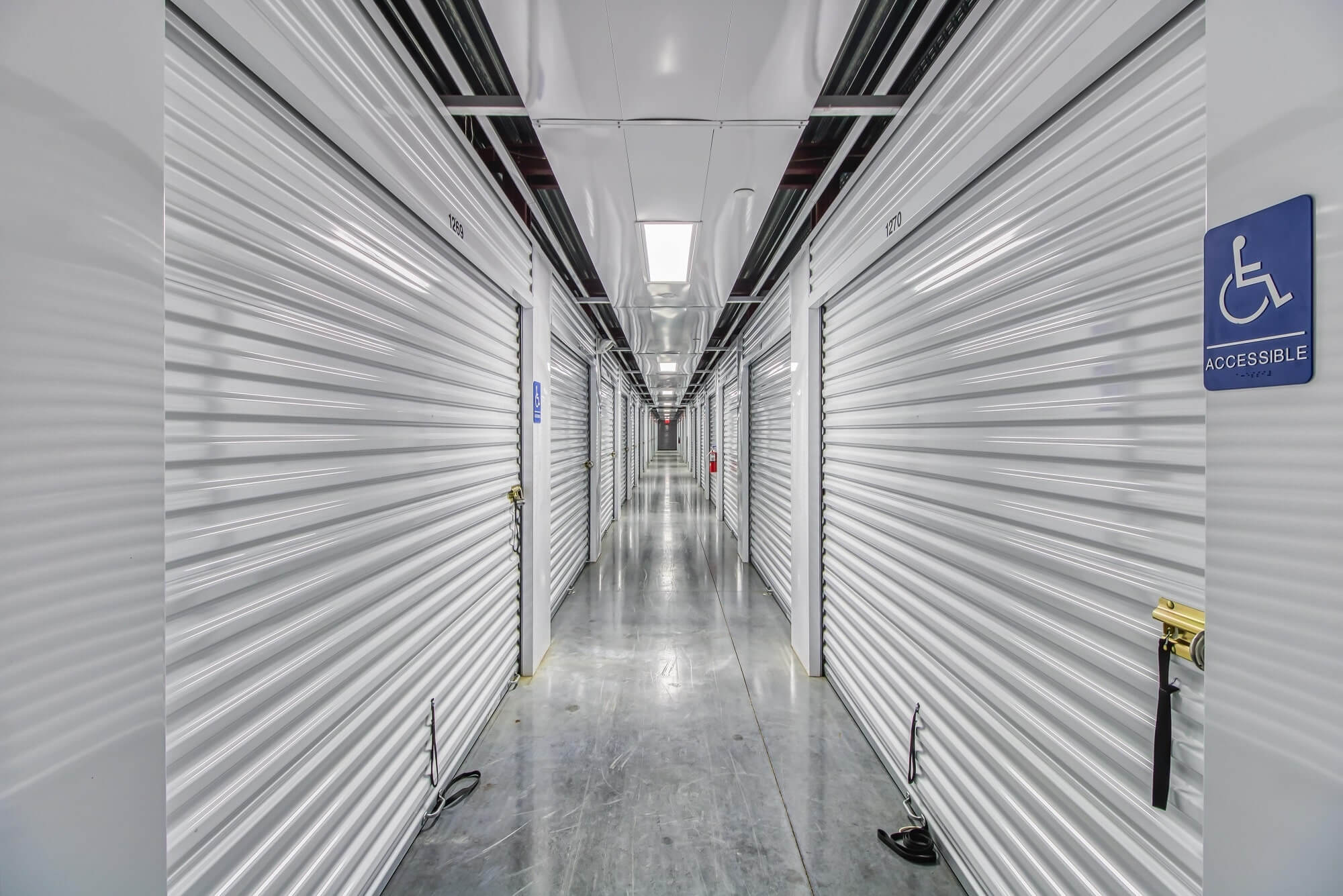 Bright hallway lined with roll-up storage doors, accessible unit signage, and polished concrete floors.