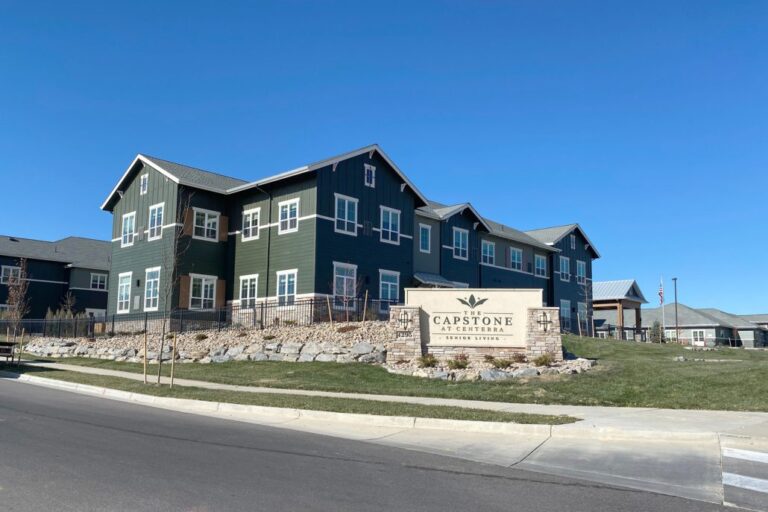 Large two-story dark green building with white-trimmed windows and gabled roofs, featuring a stone sign reading 'The Capstone at Centerra Senior Living' along the landscaped roadside.